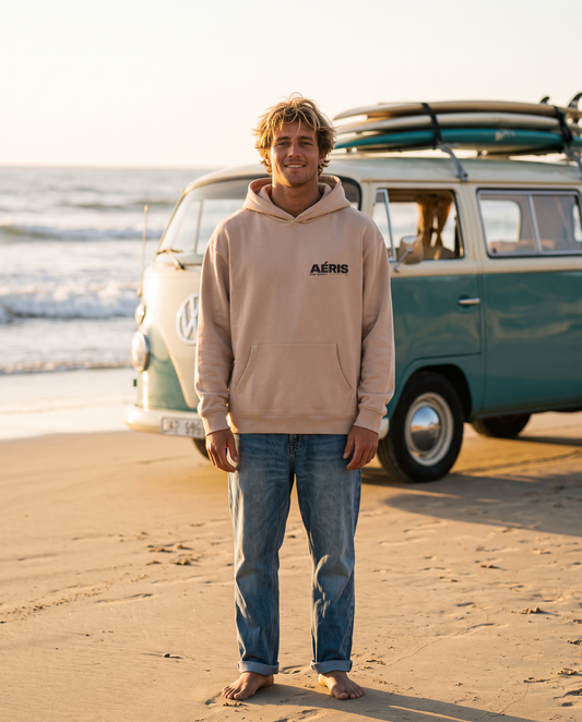 Άντρας στην παραλία φορά το oversized AέRis Surf Society hoodie σε μπεζ, μπροστά σε παλιό βανάκι με σανίδες